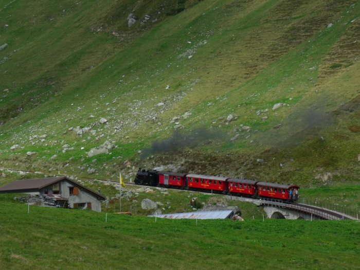 B.F.D.&nbsp;9 mit Zug Nr.133 nach Oberwald, direkt hinter dem Steinstafelviadukt, links der kleine Alm-Hof, von wo in den Sommermonaten die im Tal überall grasenden Rinder überwacht bzw. gehütet werden. Im Winter ist das alles hier unter meterhohem Schnee begraben und daher natürlich kein Betrieb, auch nicht auf dem kleinen Bauernhof, um 10:47h am 25.08.2017
